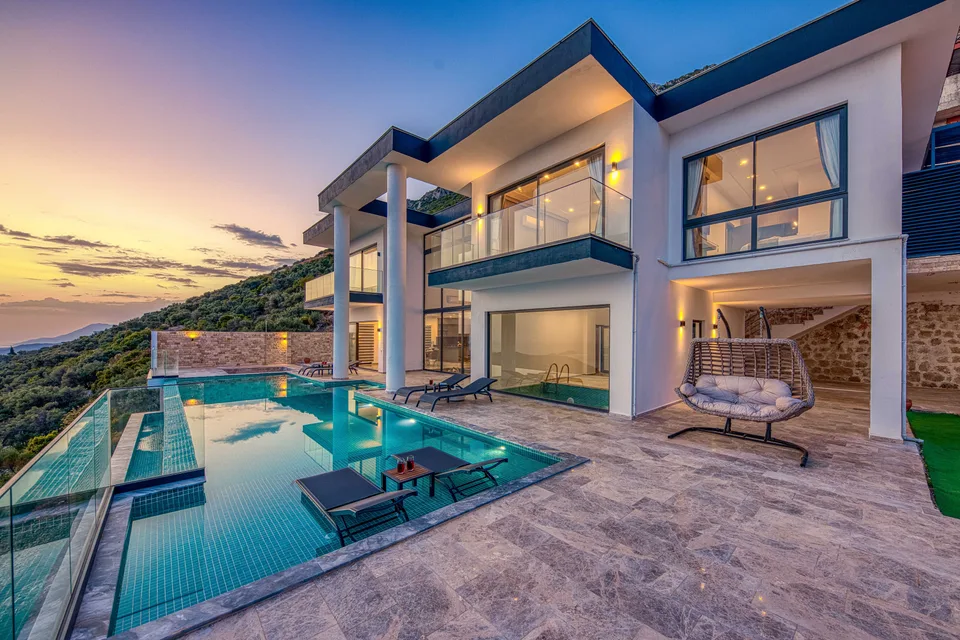 A modern villa above its infinity pool at sunset, hills and sea behind