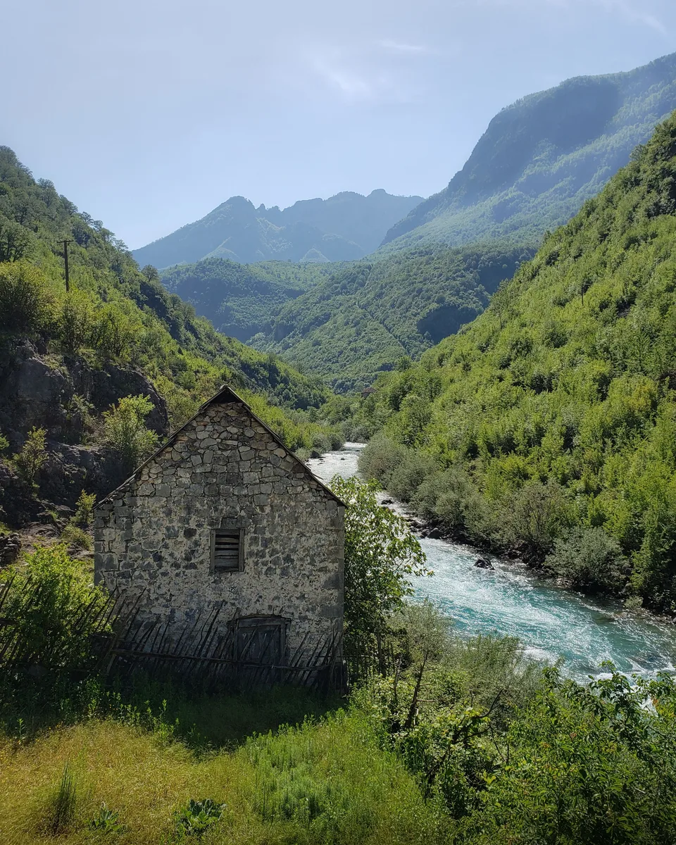 An old stone mill house on the bank of a fast mountain river, green ridges rising behind it
